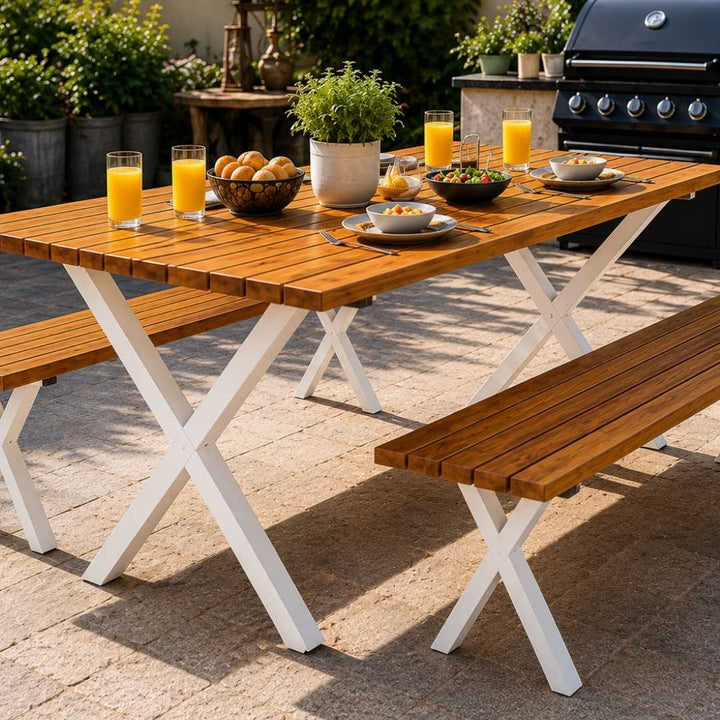 Outdoor wooden dining table with matching bench seats, shown in a patio setting with drinks, food, and a grill in the background.