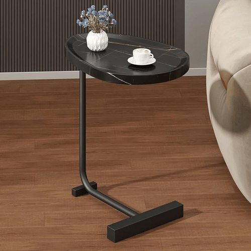 A black tear drop shaped end table with a marble top, positioned under a couch with a cup and a vase on top, set against a wood floor background.